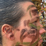 Close-up side profile of a person outdoors with camouflage face paint in brown and green on the forehead, cheek, and jawline.