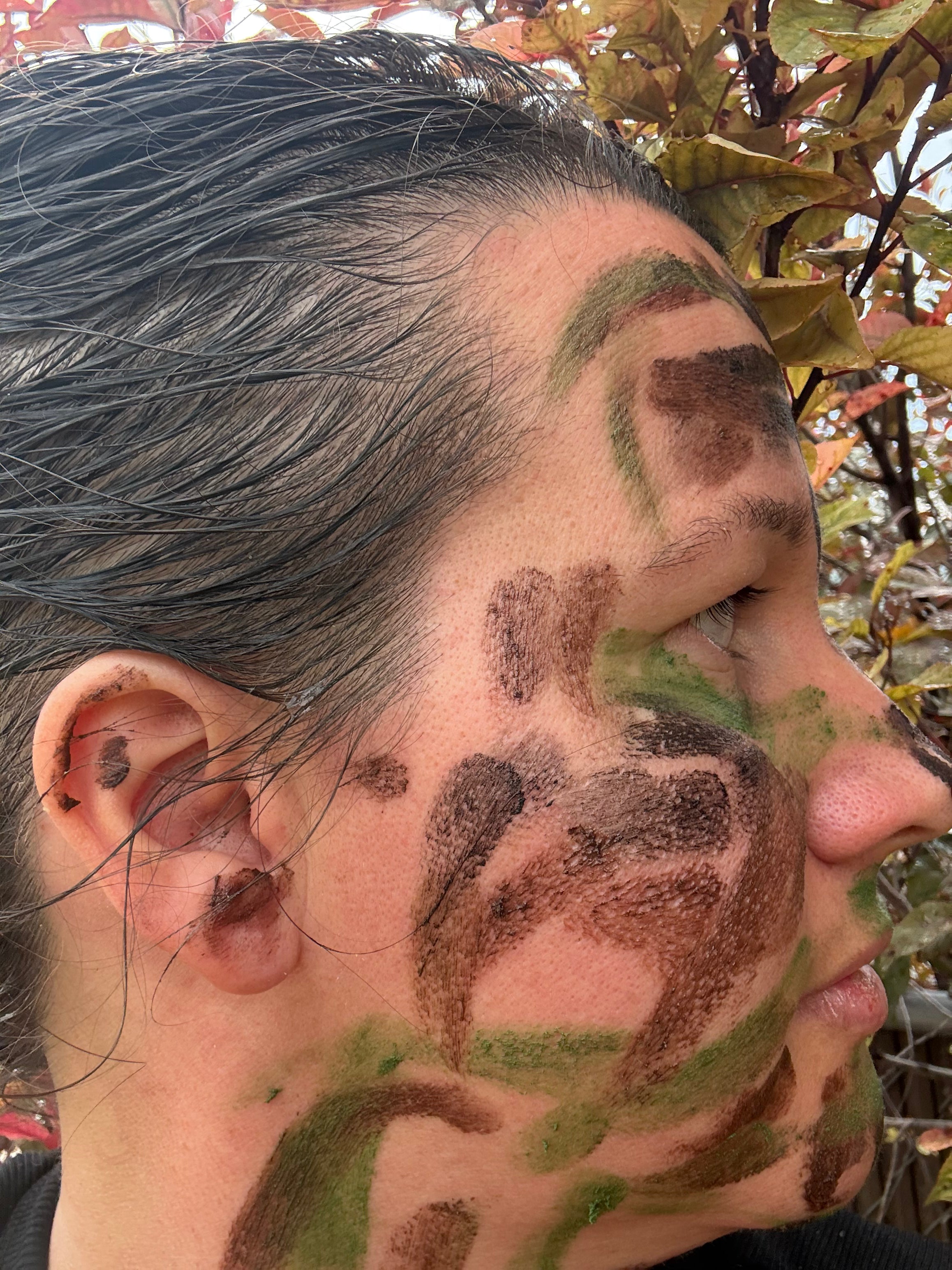 Close-up side profile of a person outdoors with camouflage face paint in brown and green on the forehead, cheek, and jawline.