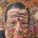 A close-up selfie of a person wearing camouflage face paint in green, brown, and black, with autumn foliage in the background.