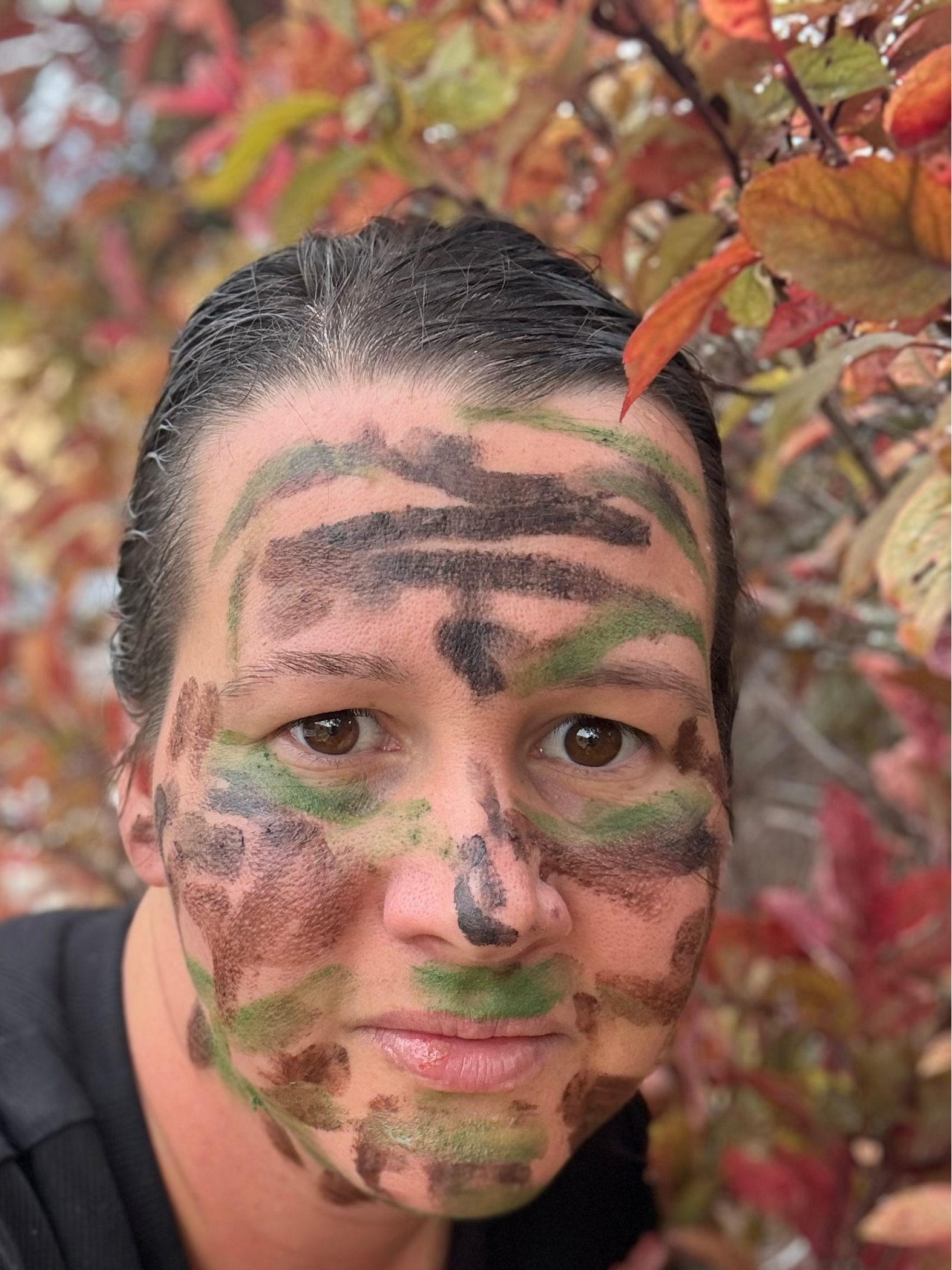 A close-up selfie of a person wearing camouflage face paint in green, brown, and black, with autumn foliage in the background.