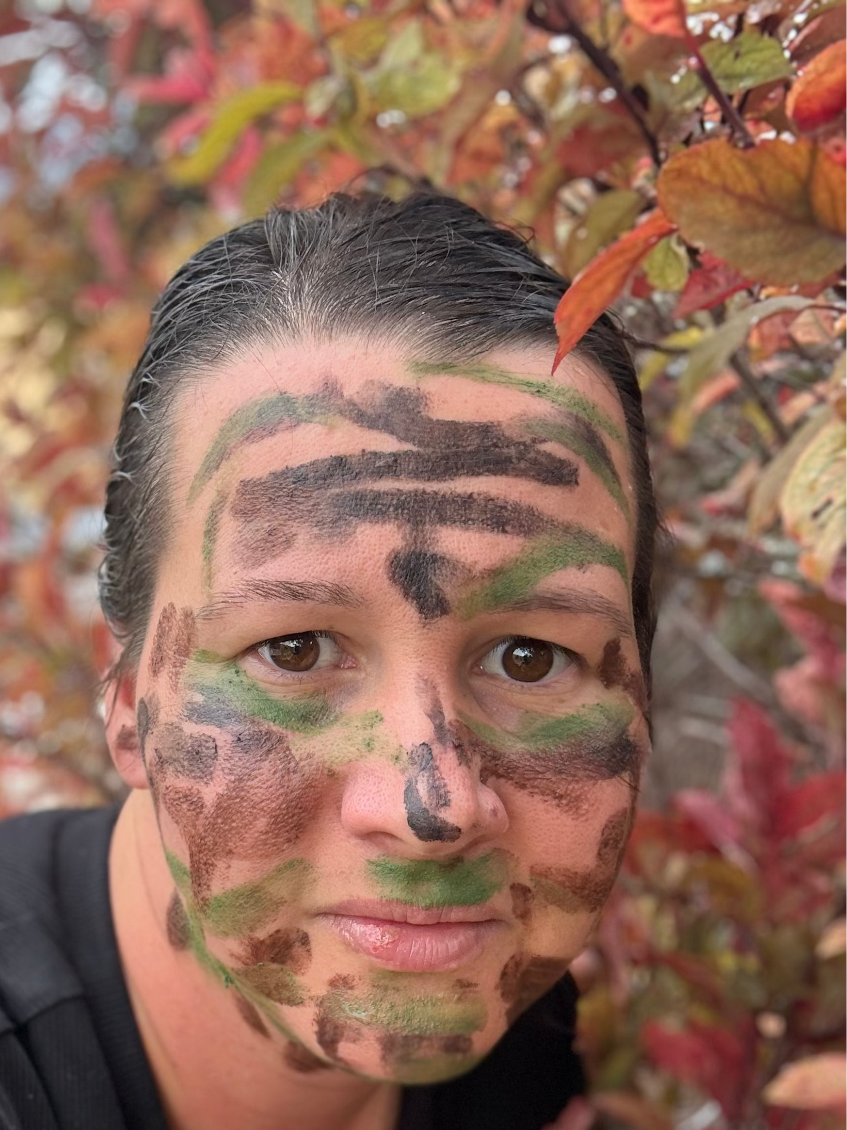 A close-up selfie of a person wearing camouflage face paint in green, brown, and black, with autumn foliage in the background.
