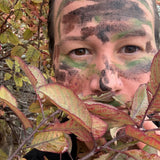 A close-up outdoor portrait of a person with camouflage face paint in green, black, and brown, partially obscured by red and green leaves in the foreground.