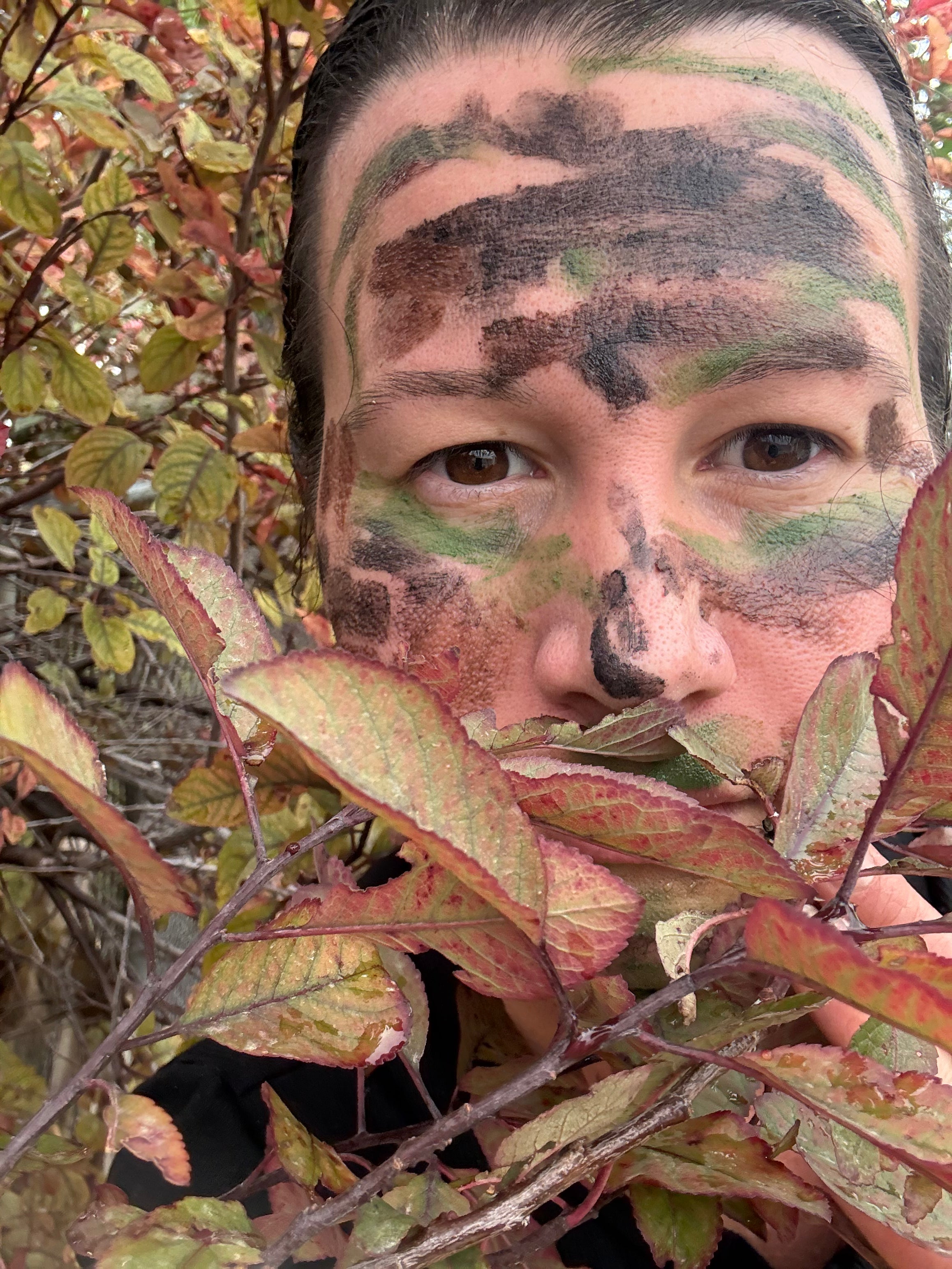 A close-up outdoor portrait of a person with camouflage face paint in green, black, and brown, partially obscured by red and green leaves in the foreground.