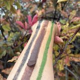 A close-up of a forearm with three painted color bands: a dark brown stripe, a green stripe, and a black stripe, shown outdoors against blurred autumn leaves.