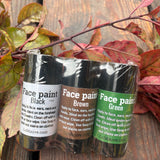 Three rolls of face paint in black, brown, and green, packaged together in a clear plastic wrap on a rustic wooden surface with autumn leaves.