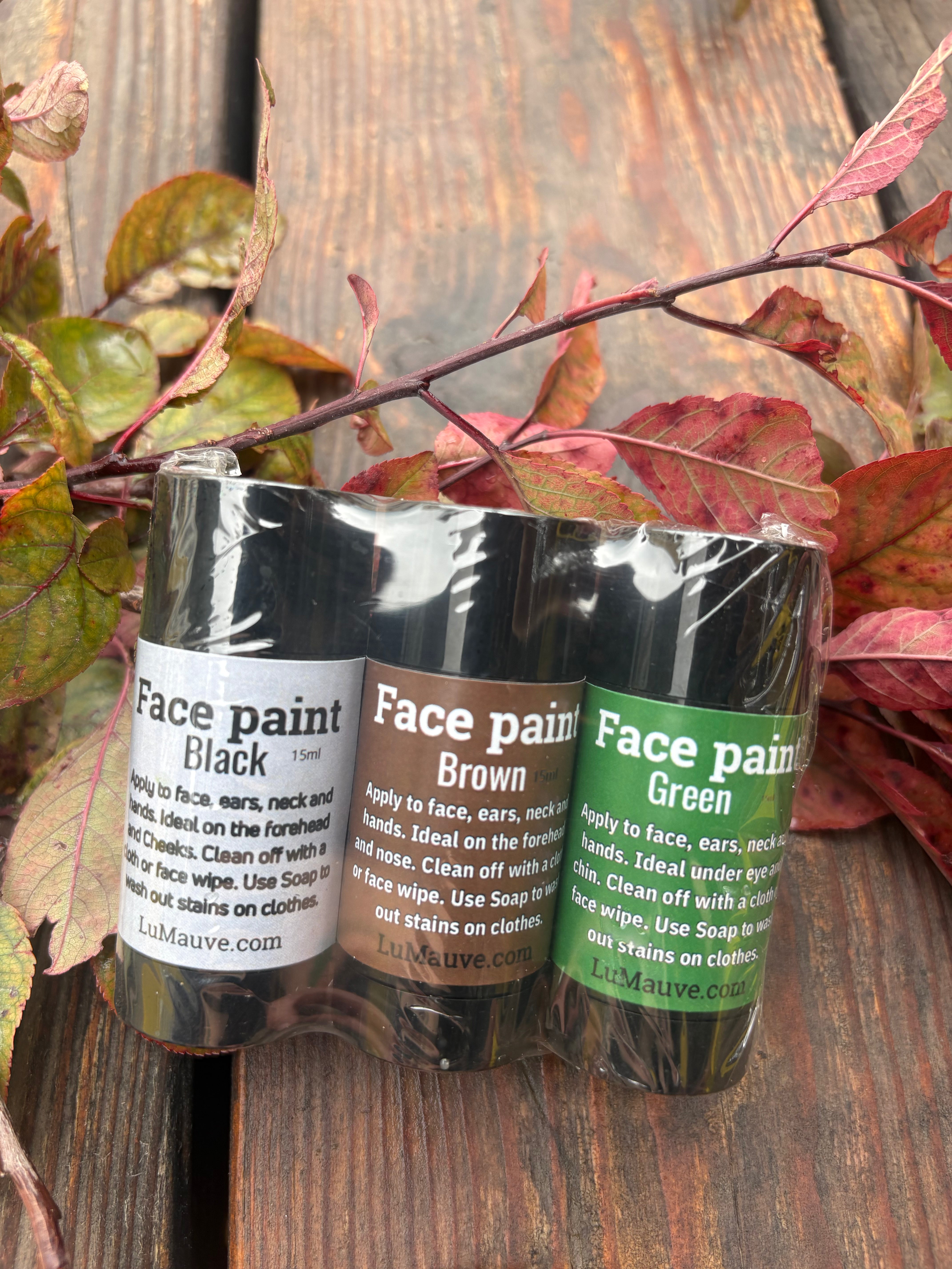 Three rolls of face paint in black, brown, and green, packaged together in a clear plastic wrap on a rustic wooden surface with autumn leaves.