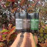 Three tubes of face paint in black, brown, and green shades are shown on a person's forearm, with a background of red and green autumn leaves.
