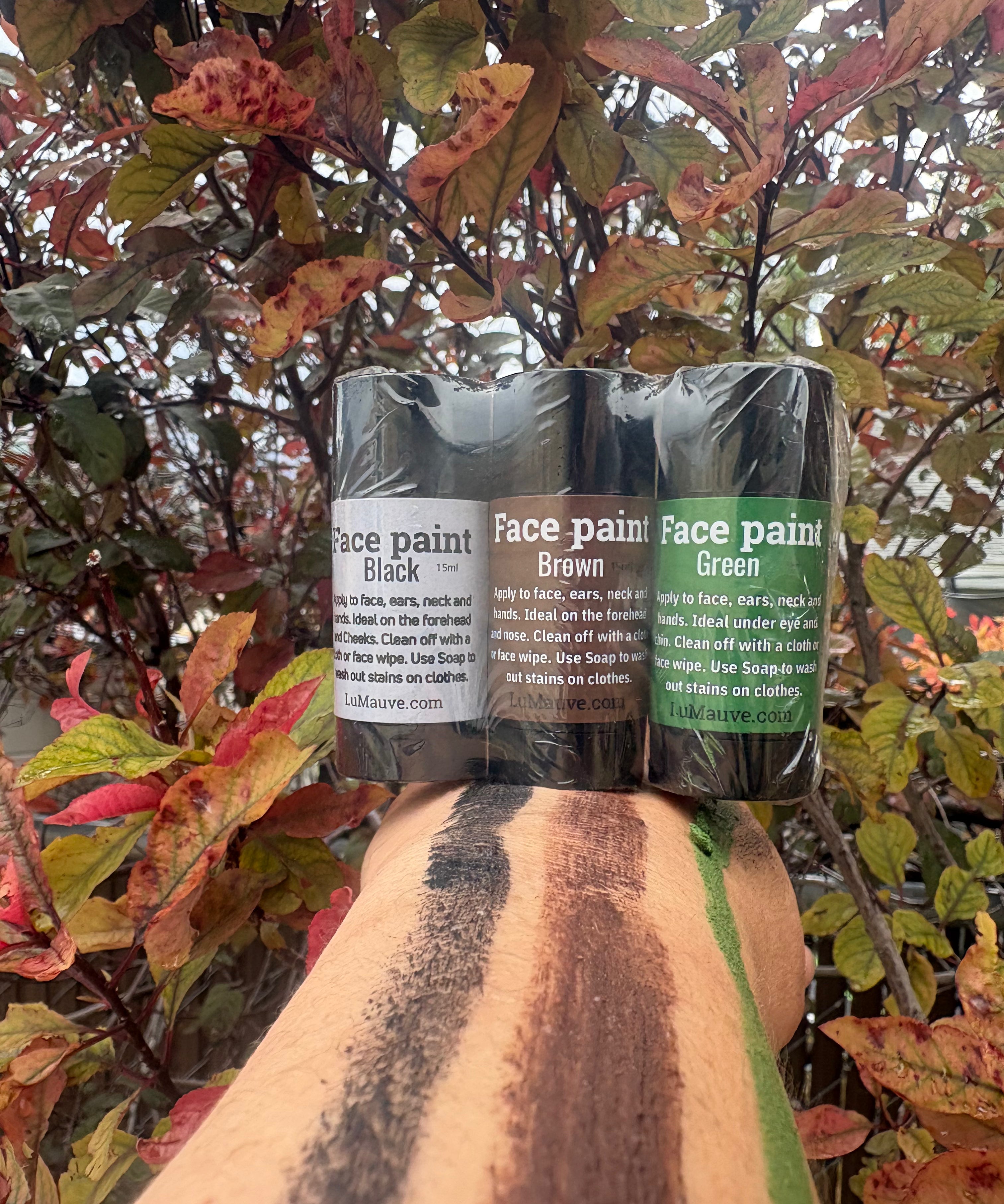 Three tubes of face paint in black, brown, and green shades are shown on a person's forearm, with a background of red and green autumn leaves.