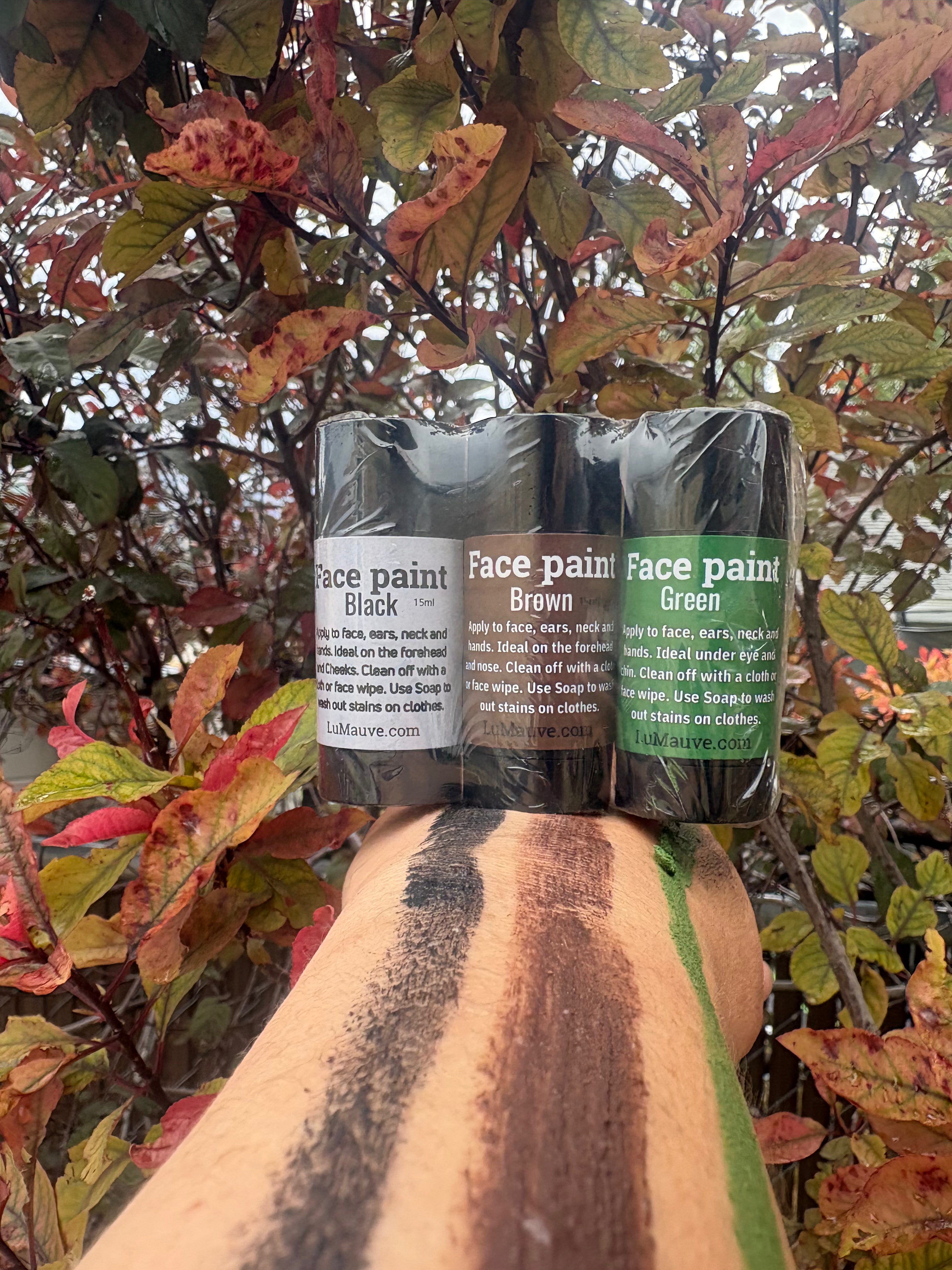 Three tubes of face paint in black, brown, and green shades are shown on a person's forearm, with a background of red and green autumn leaves.