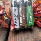 Three face paint tubes in black, brown, and green colors, packaged together in a clear plastic sleeve and resting on a rustic wooden surface surrounded by red autumn leaves.