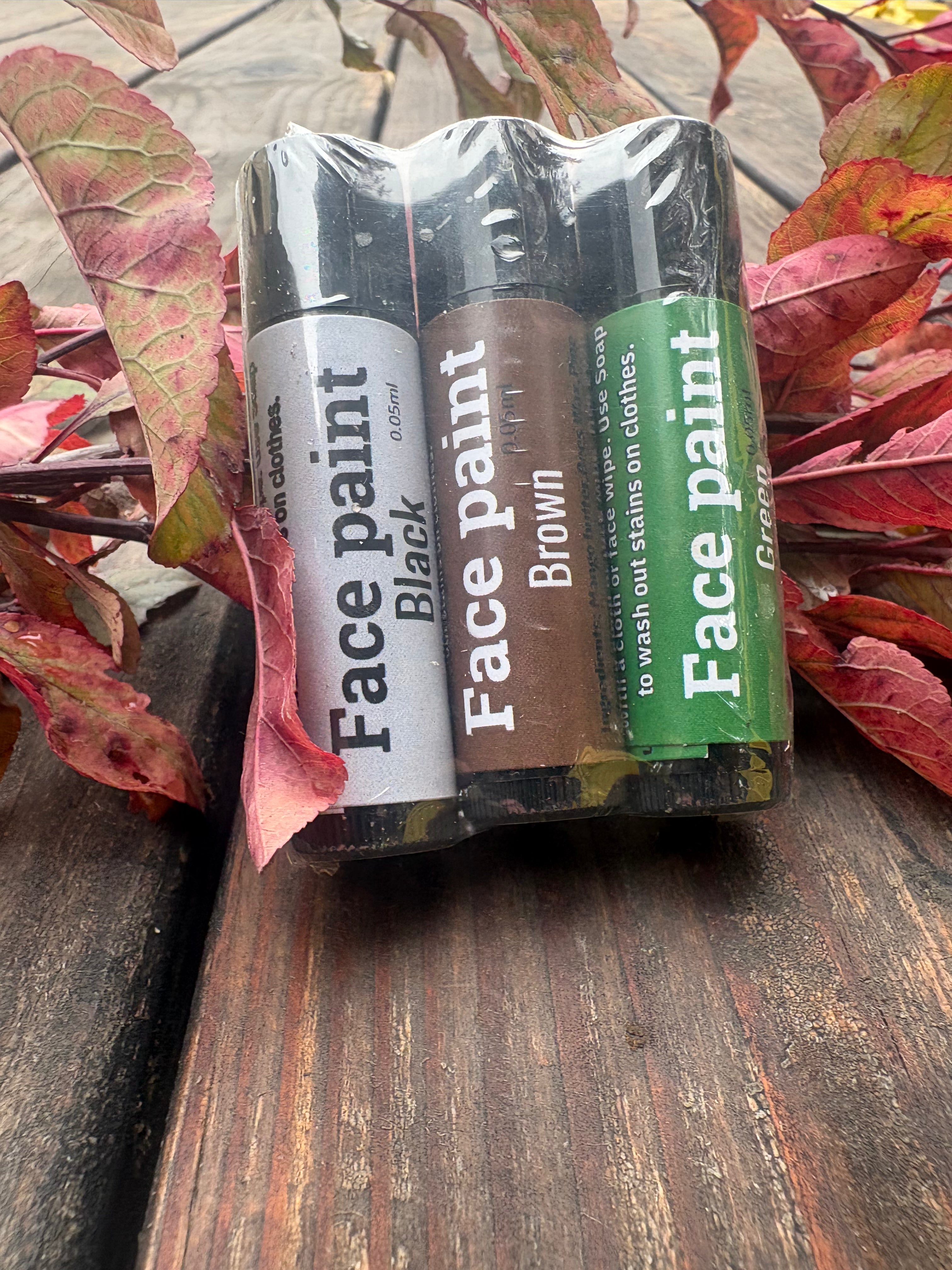 Three face paint tubes in black, brown, and green colors, packaged together in a clear plastic sleeve and resting on a rustic wooden surface surrounded by red autumn leaves.