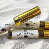 Two gold and white cosmetic tubes labeled 'Lash & Brow' on a textured white surface.