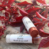 Two lip balm tubes labeled 'Rose Blush & Lip Color' on a wooden surface with red leaves in the background.