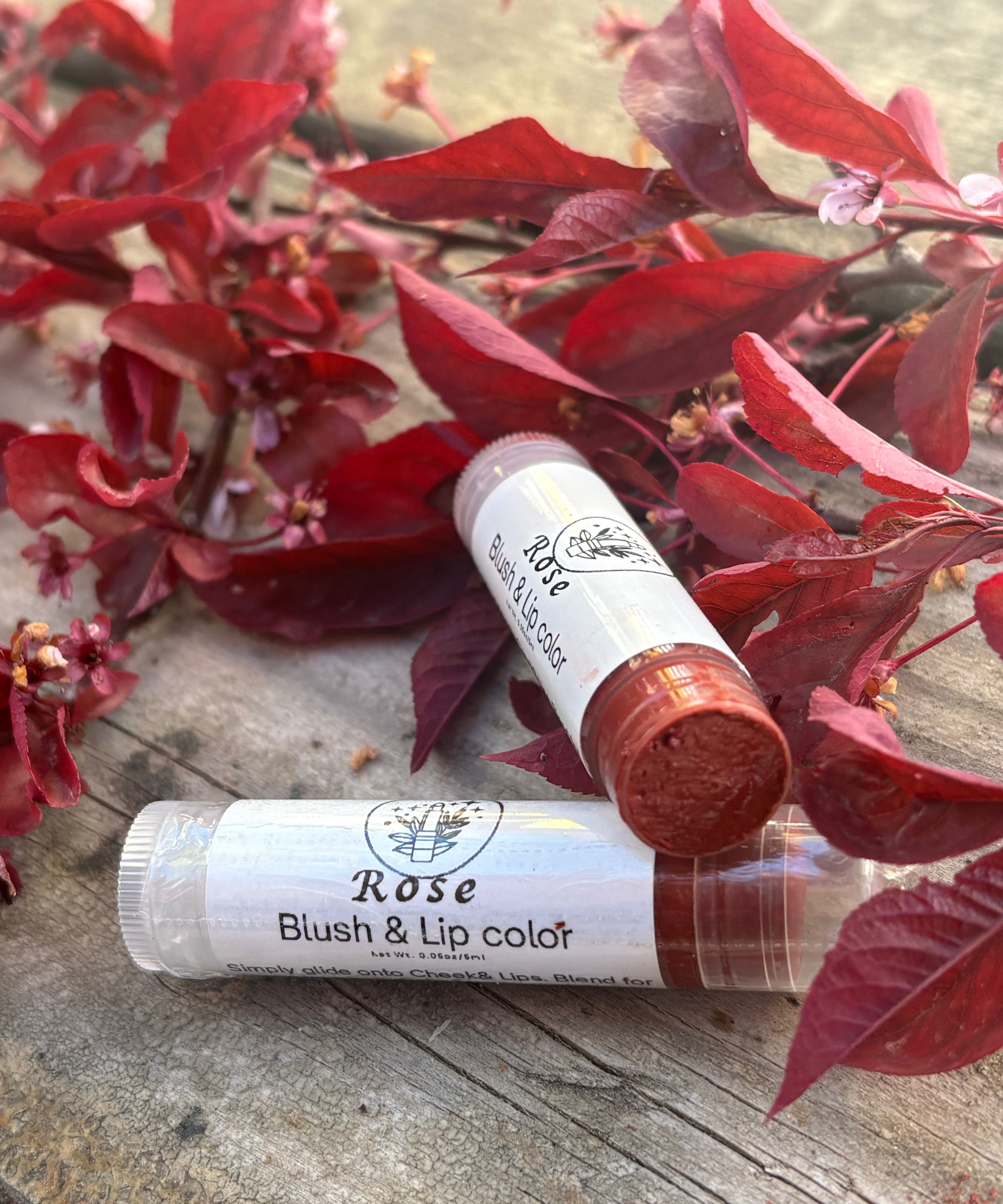 Two lip balm tubes labeled 'Rose Blush & Lip Color' on a wooden surface with red leaves in the background.