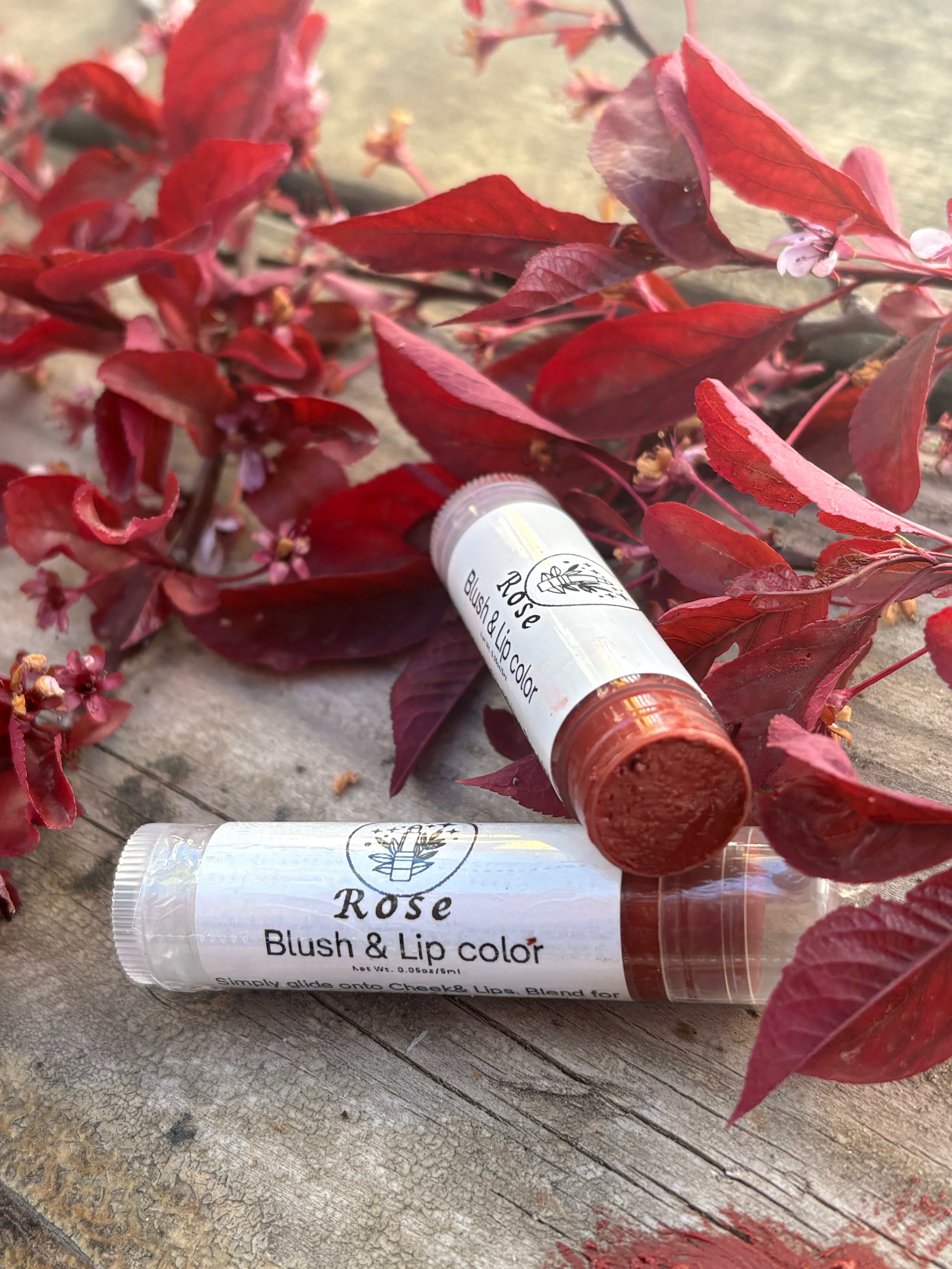 Two lip balm tubes labeled 'Rose Blush & Lip Color' on a wooden surface with red leaves in the background.