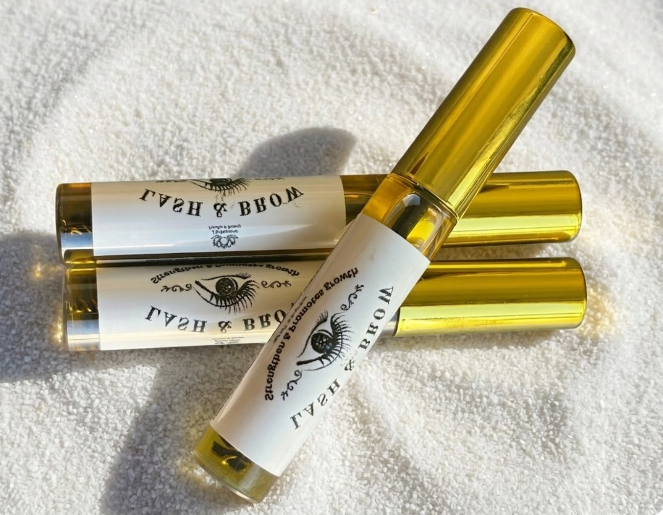 Three gold lash bottles with labels on a textured white surface